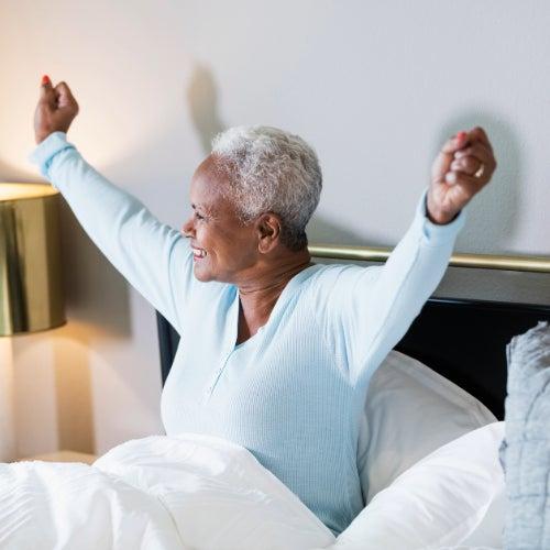 A woman with short white hair in blue pajamas stretches her arms in bed and smiles