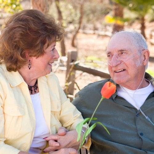 An older man with a portable oxygen tube in his nose sits on a bench outdoors and talks to a woman