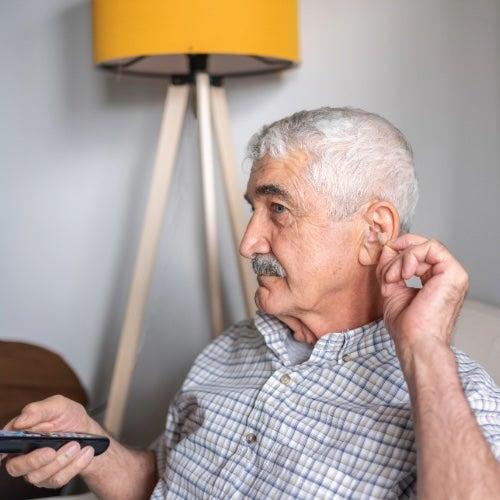 A man with grey hair puts in a hearing aid while holding a remote control
