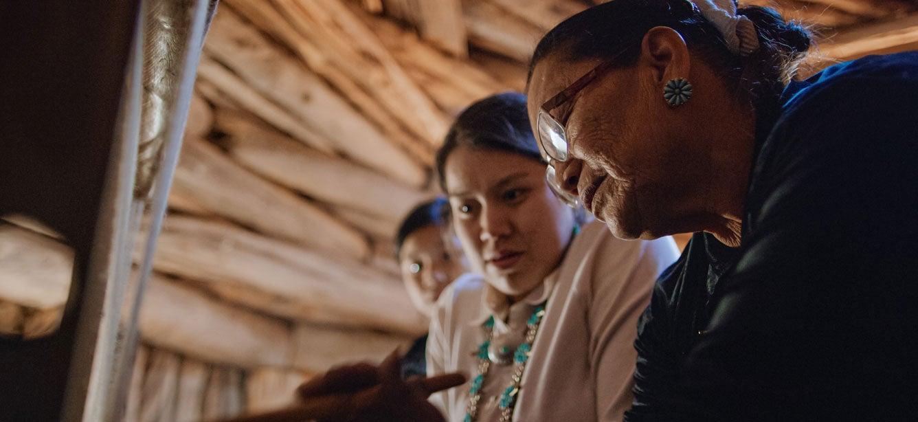 A senior Native American woman is showing a younger generation how to weave.