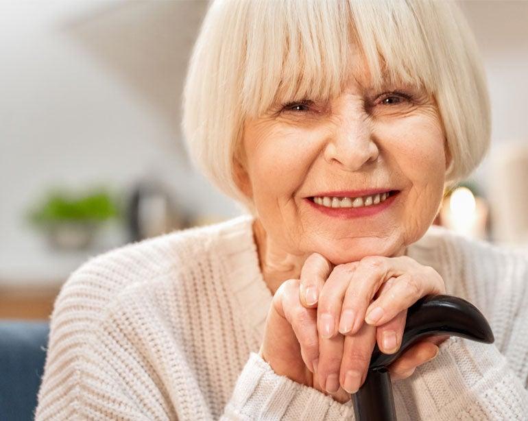 An up close shot of a senior woman smiling at the camera with her hands resting on her cane.