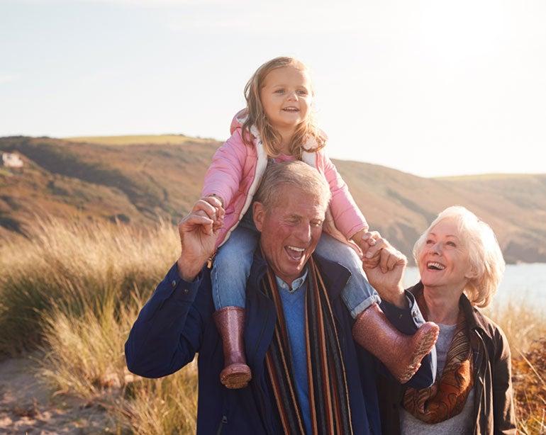 An older adult grandfather has his granddaughter on his shoulders while his wife looks at both of them lovingly.