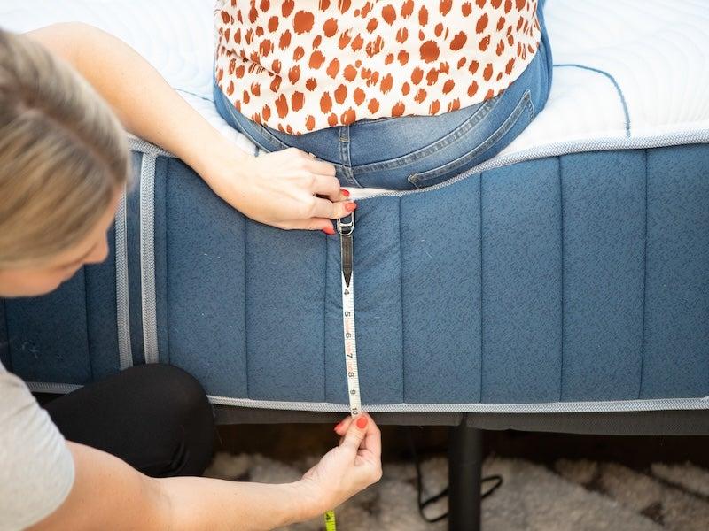 A woman measures how far the Bear Elite Hybrid mattress compresses when a tester sits on the edge.