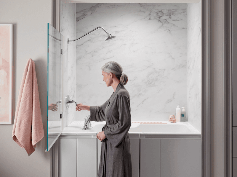 A woman in a gray robe adjusts the shower knob in a modern bathroom with marble walls. A soft pink towel hangs nearby, creating a calm, serene setting.