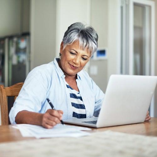 A woman sitting at a table with an open laptop writing notes on paper