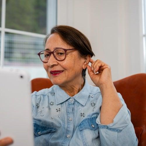 A woman with short black hair and glasses adjusts her hearing aid while looking at a phone