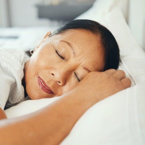 A woman with black hair lies on a white pillow and smiles with her eyes closed