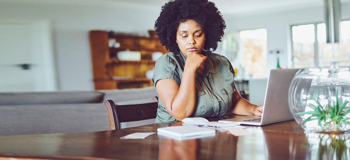 Woman with curly hair sits at a table, gazing at a laptop, with papers, a notebook, and a plant in a glass bowl nearby.