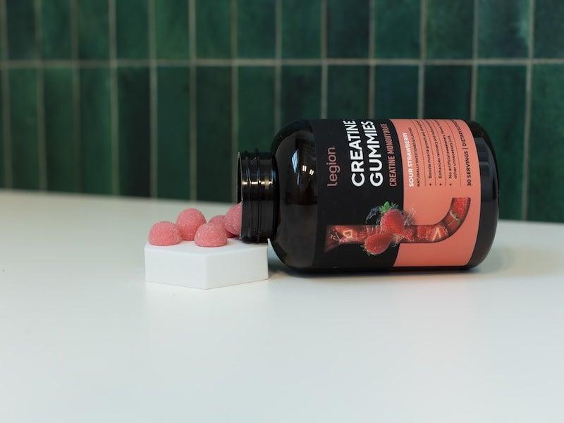 Legion Creatine Gummies bottle on its side, spilling little pink gumdrops onto a hexagonal pedestal