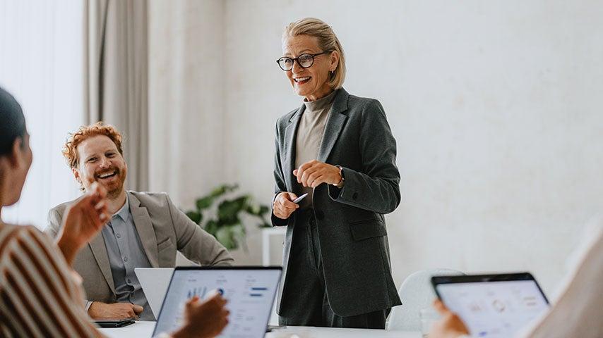 smiling middle aged woman standing and talking with colleagues seated at laptops
