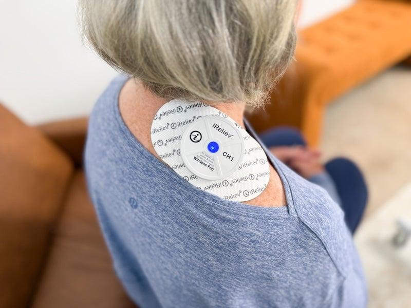A woman with a heather-blue long-sleeve shirt wears an iReliev electrode pad on the back of her neck