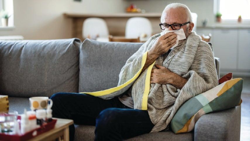 A senior man wearing black glasses is sitting on his couch wrapped in a blanket and blowing his nose.