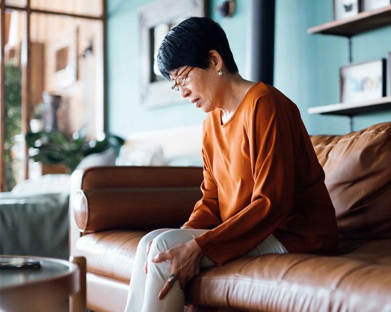 An older woman with short black hair and glasses sits on a brown leather sofa in a living room, looking down at her knee with a pained expression, possibly indicating bone pain. She is wearing an orange long-sleeve top and light-colored pants. The setting is cozy, with soft lighting and a few decorative items visible in the background. The woman appears to be experiencing discomfort or pain in her knee.