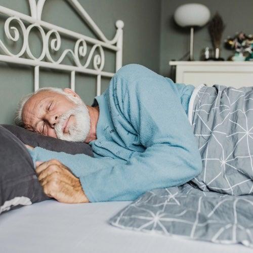 An older man in blue pajamas sleeps on a bed on a white metal frame bed