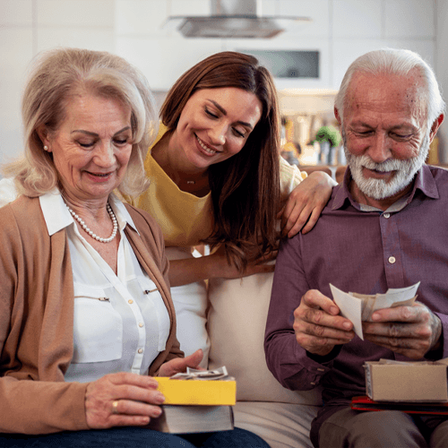 A woman and a man are sitting on a couch, opening gifts while another woman looks over their shoulders and smiles