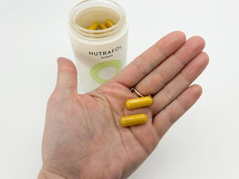 An open container of Nutrafol supplements and two capsules in a person's hand