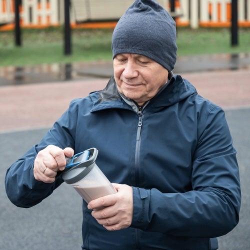 An older man stands in an outdoor workout area and opens a protein drink