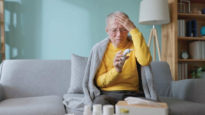 A senior man in a yellow long-sleeve shirt is taking his temperature while wrapped in a blanket on his couch.