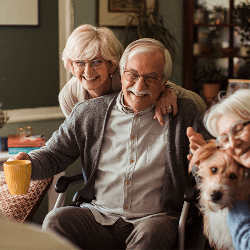 Three older adults huddle together. The man in the center holds a yellow mug, and the woman on the right holds a dog. All three people in the photo have glasses.