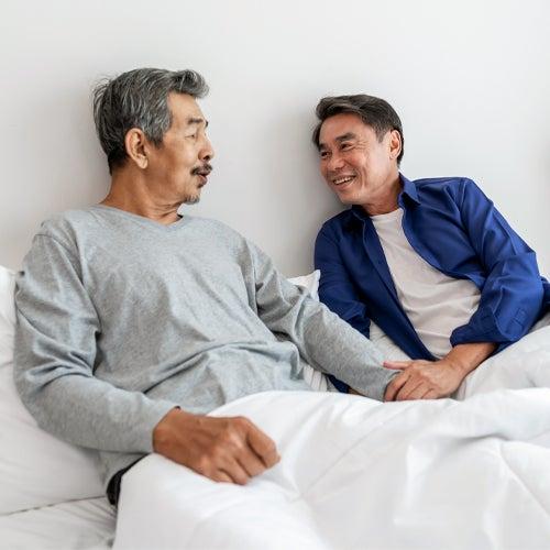 Two men smile while sitting up in a bed with white sheets