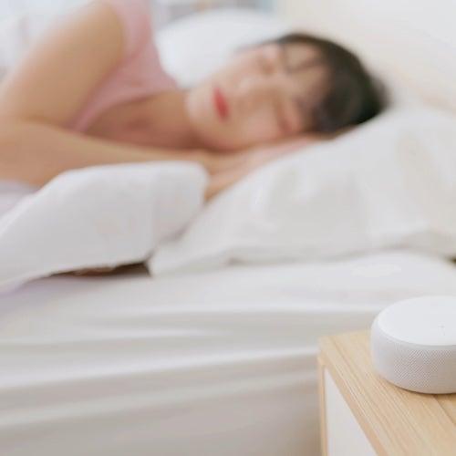 A woman rests on her side in bed next to a table holding a white noise machine in the foreground