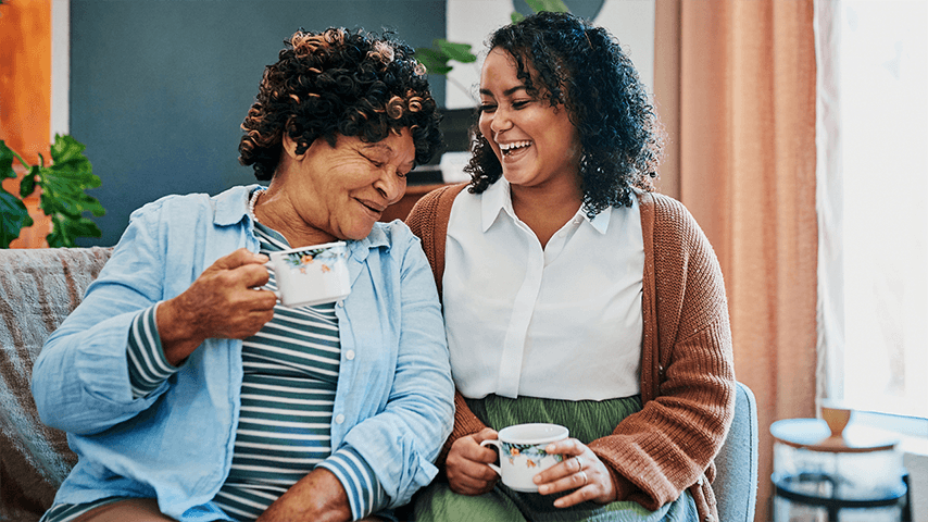 Two women, one older and one younger, sitting on a couch, smiling and enjoying a cup of tea together in a cozy living room
