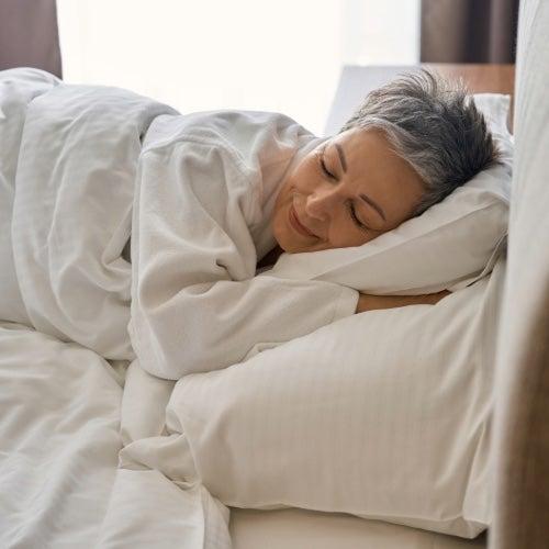 A woman with short, white hair sleeps in a comfortable-looking bed with white sheets