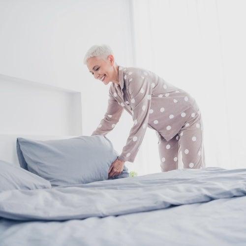 A woman with short white hair in polka dot pajamas fluffs up a pillow on a grey bed