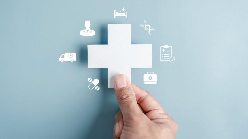 A hand holds up a medical cross symbol, surrounded by other medical icons, including medication, a doctor's bag, a clipboard, a DNA strand, a hospital bed, a physician, and an ambulance.