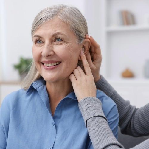 A woman in a blue shirt with grey hair smiles as her friend helps position a hearing aid in her ear