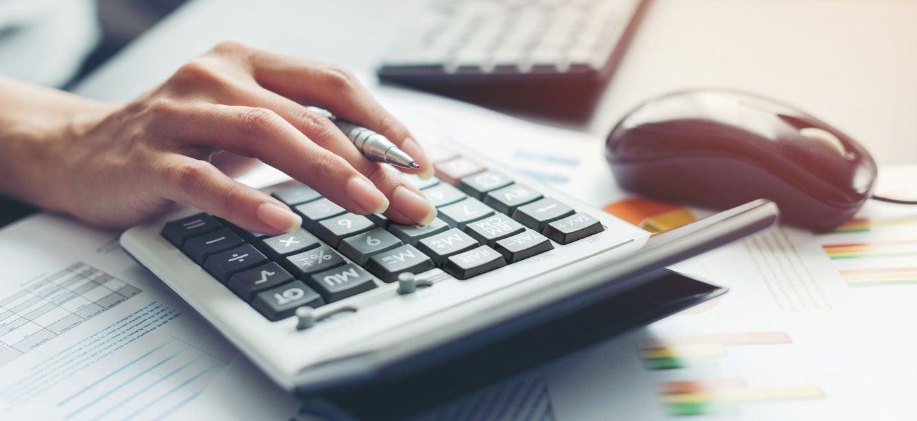 Hand pressing buttons on a calculator, with papers and a black computer mouse in the background.