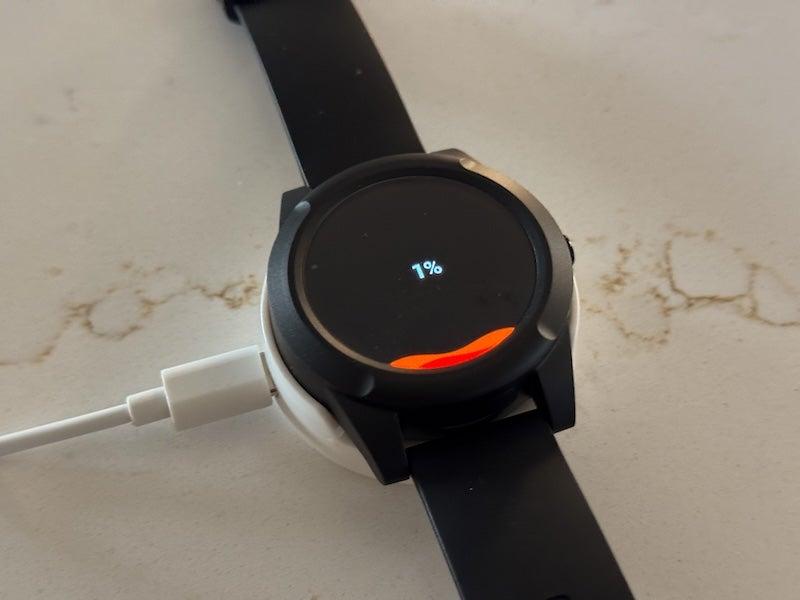 A circular black digital watch face displays a wavelike red waterline near the bottom of the screen that shows the battery charge level. Above the waterline, the display reads “1%.” The black watch sits on a white charging cradle; a white cord pokes out of the cradle’s left side.