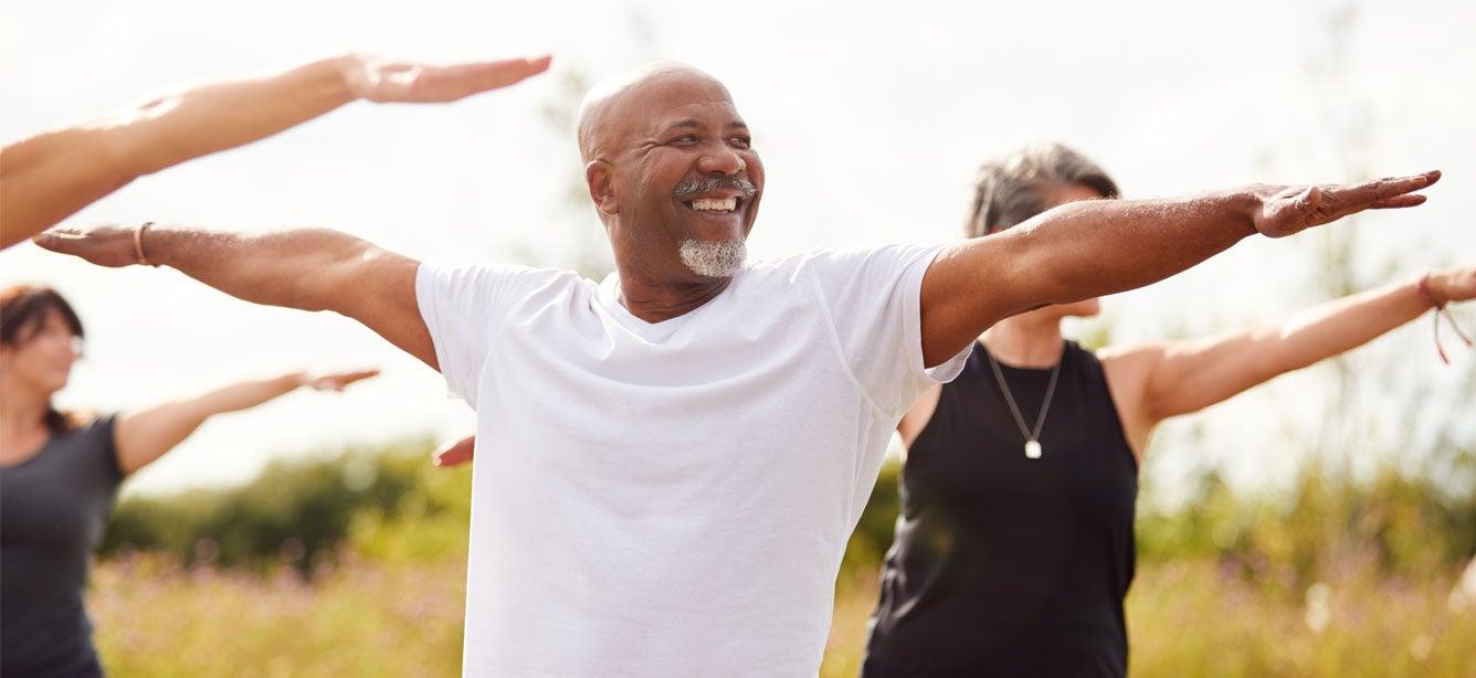 A Black senior man is practicing yoga in the park with other older adults.
