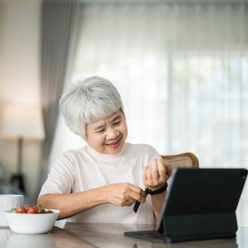 A woman sitting at a table in front of a tablet device putting on smart health watch
