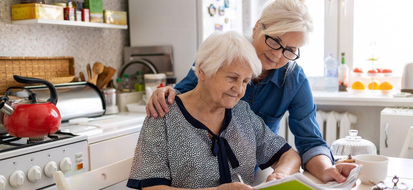 A mature woman looks over her elderly mother's shoulder while helping her with paperwork.