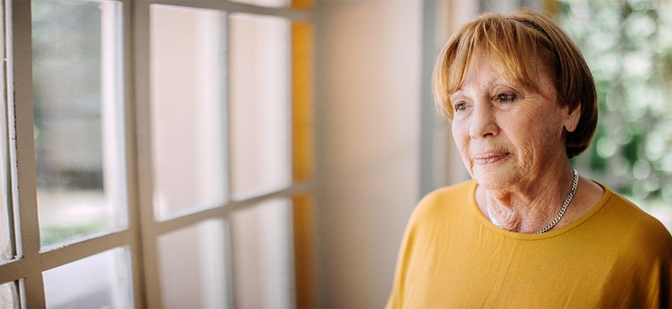 A senior woman sad and alone stares out her window, feeling socially isolated from the pandemic.
