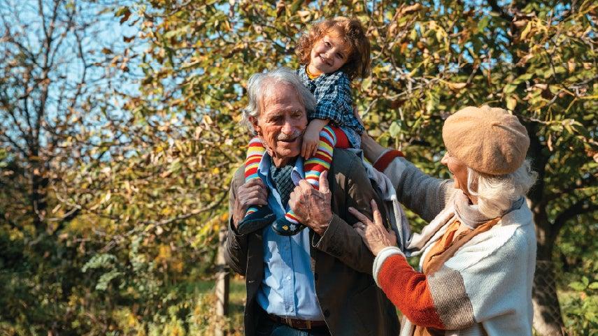 Caucasian grandparents are outdoors in the fall walking with their grandchild. The grandfather has the young child on his shoulders.