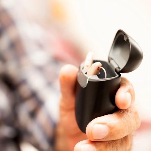 A hand holds a pair of hearing aids in their charger