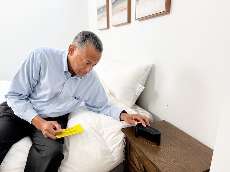 A man in a periwinkle button-down shirt sits on a bed. He holds a yellow instruction booklet and adjusts the buttons on an all-black alarm clock
