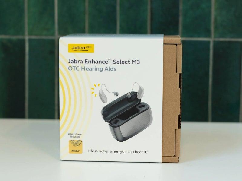 The Jabra Enhance Select M3 hearing aids in their box in front of a green tile background