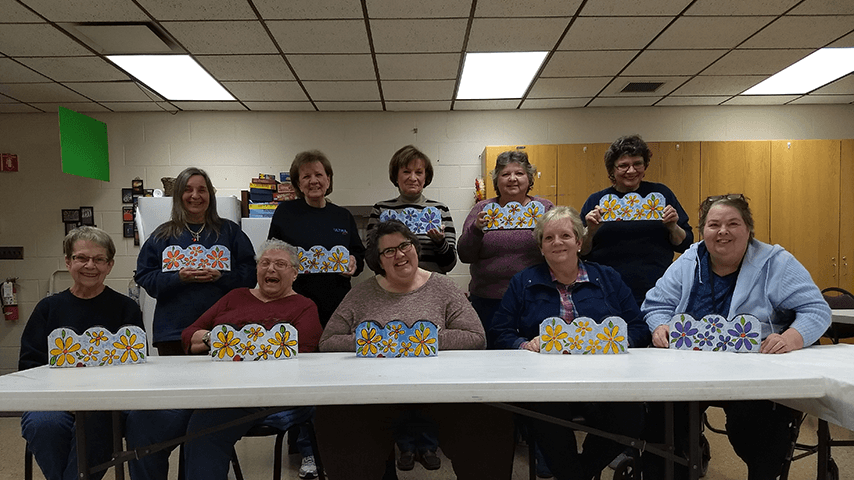 10 smiling older women showing flower crafts at senior center table