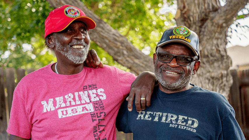 Two smiling older men wearing veteran ball caps and T-shirts stand arm in arm outside in front of a tree