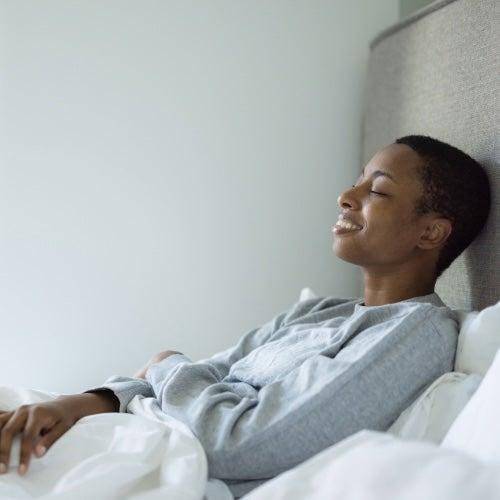 A woman in grey pajamas smiles and lies propped up on white sheets