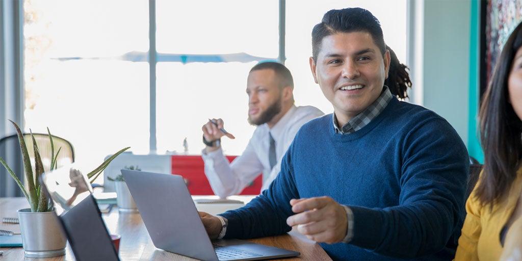 A Hispanic mail professional is setting in a meeting looking at his laptop and explaining material to another colleague.