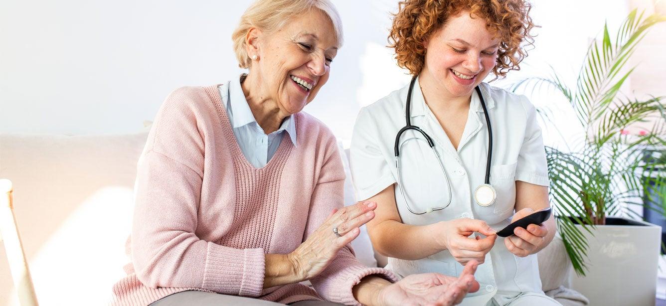 Older woman in pink sweater smiles at a healthcare provider in medical attire holding a phone.