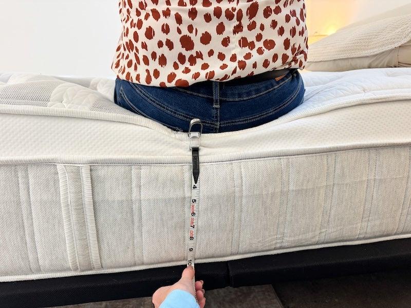 A person holds a tape measure up to the Leesa Plus hybrid mattress while another person sits on it.