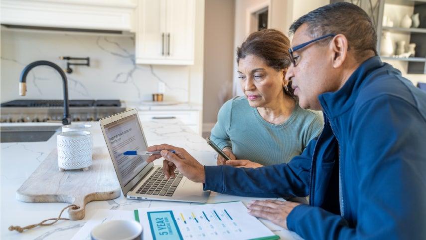 An older man is pointing his pen at the screen of his laptop while having a conversation about Medicare with a Hispanic-looking older woman.