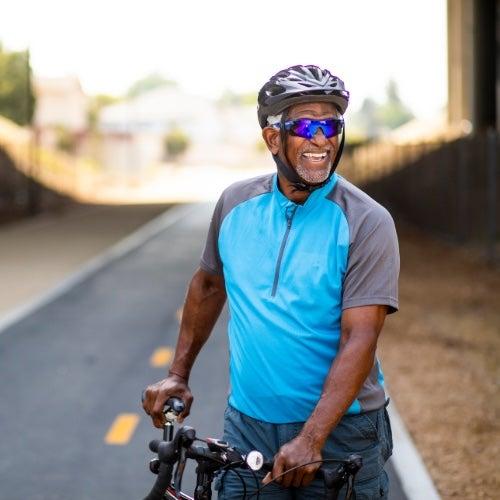 A man in shiny, wraparound sunglasses smiles while holding up his bike