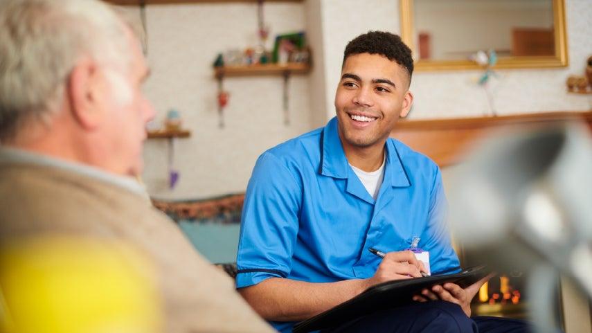 A young Black male paid caregiver is holding a clipboard while talking to a male older adult about providing his services.