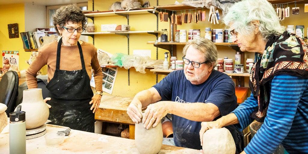 Two older women watching an older man work on pottery in an art studio